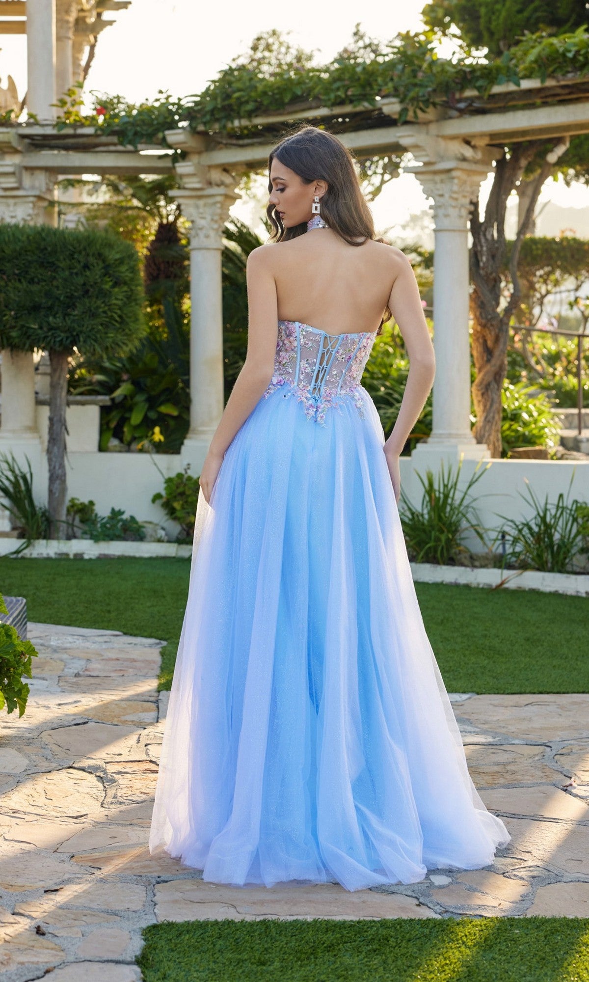 Amid lush greenery and stone columns, a woman faces away outdoors in the Long Prom Dress: Juliet JT2682A—strapless, with a glittering sheer embellished bodice and flowing tulle skirt.